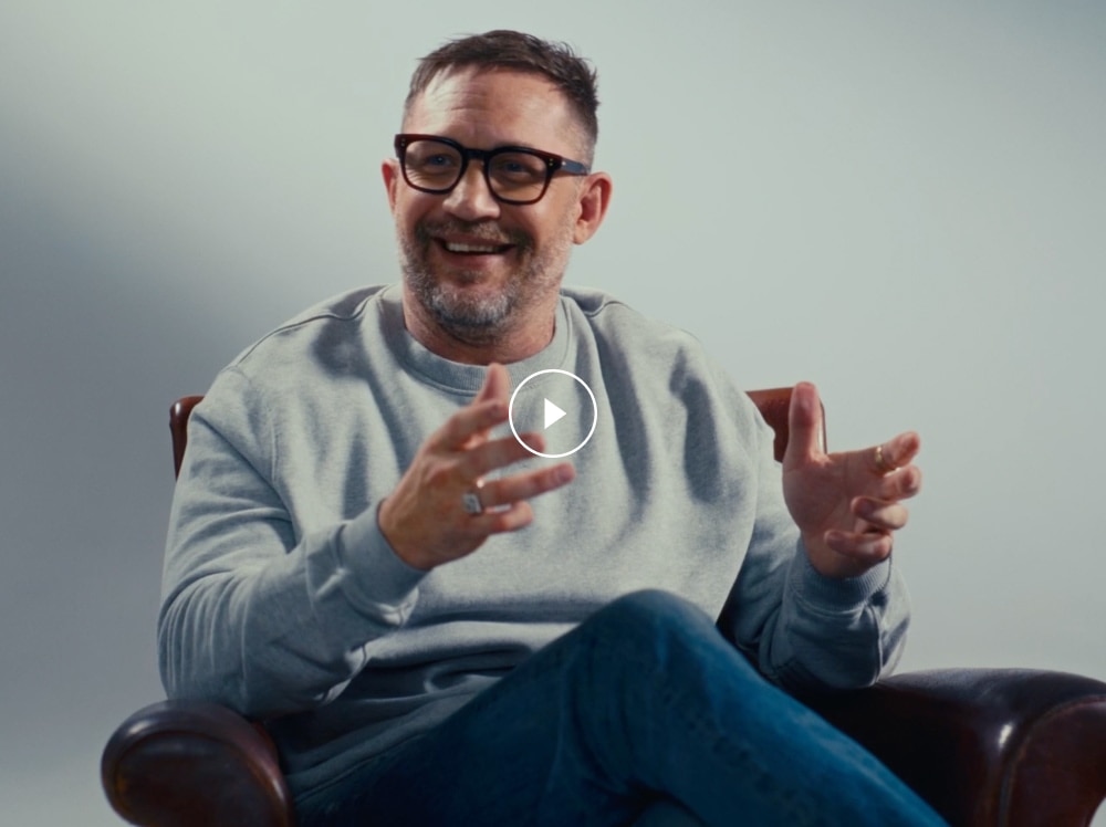 A clapperboard reveals Tom Hardy sat next to his father, against a light backdrop. He is wearing a grey jumper and glasses.