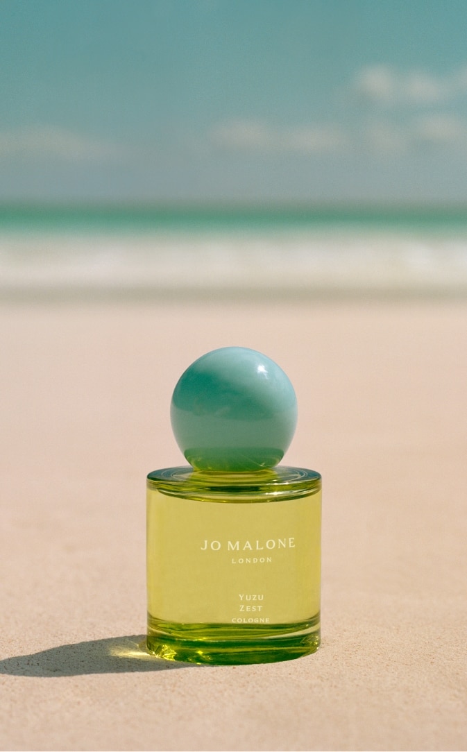 Jo Malone London Frangipani cologne sat on a tropical beach. A clear bottle with white cap.