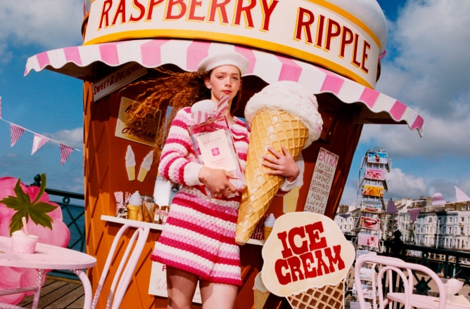 Une personne en rose tient une crème glacée et une Jo Malone London eau de Cologne sur un quai, avec des sièges roses et une hutte géante en forme de cône.