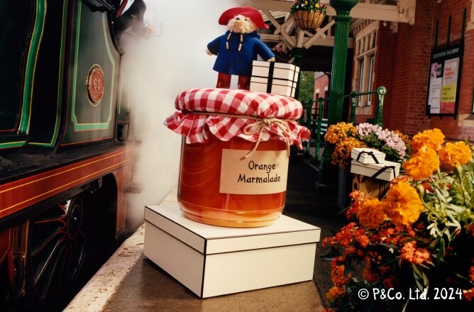 L'ours Paddington à côté de boîtes couleur crème sur un pot géant de marmelade à l'orange, accompagné d'un conducteur de train sortant d'un train en marche
