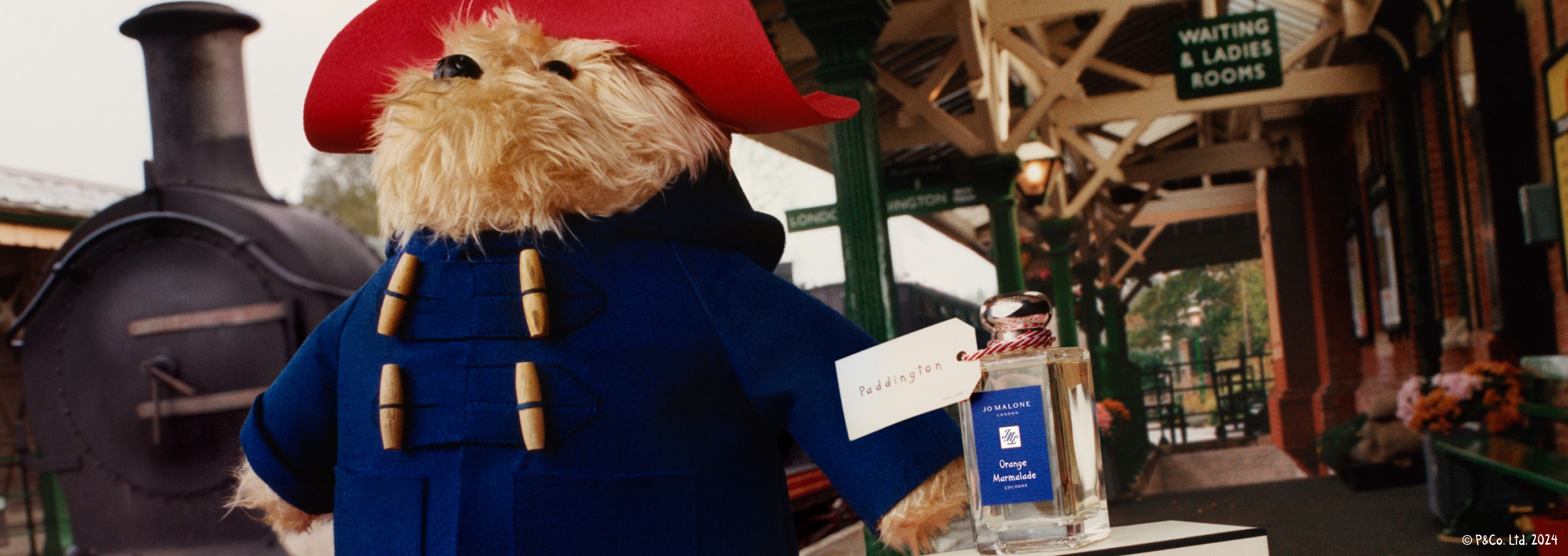 L’ours Paddington et la Cologne Orange Marmalade avec une étiquette Paddington accrochée au flacon en verre transparent dans une gare. 